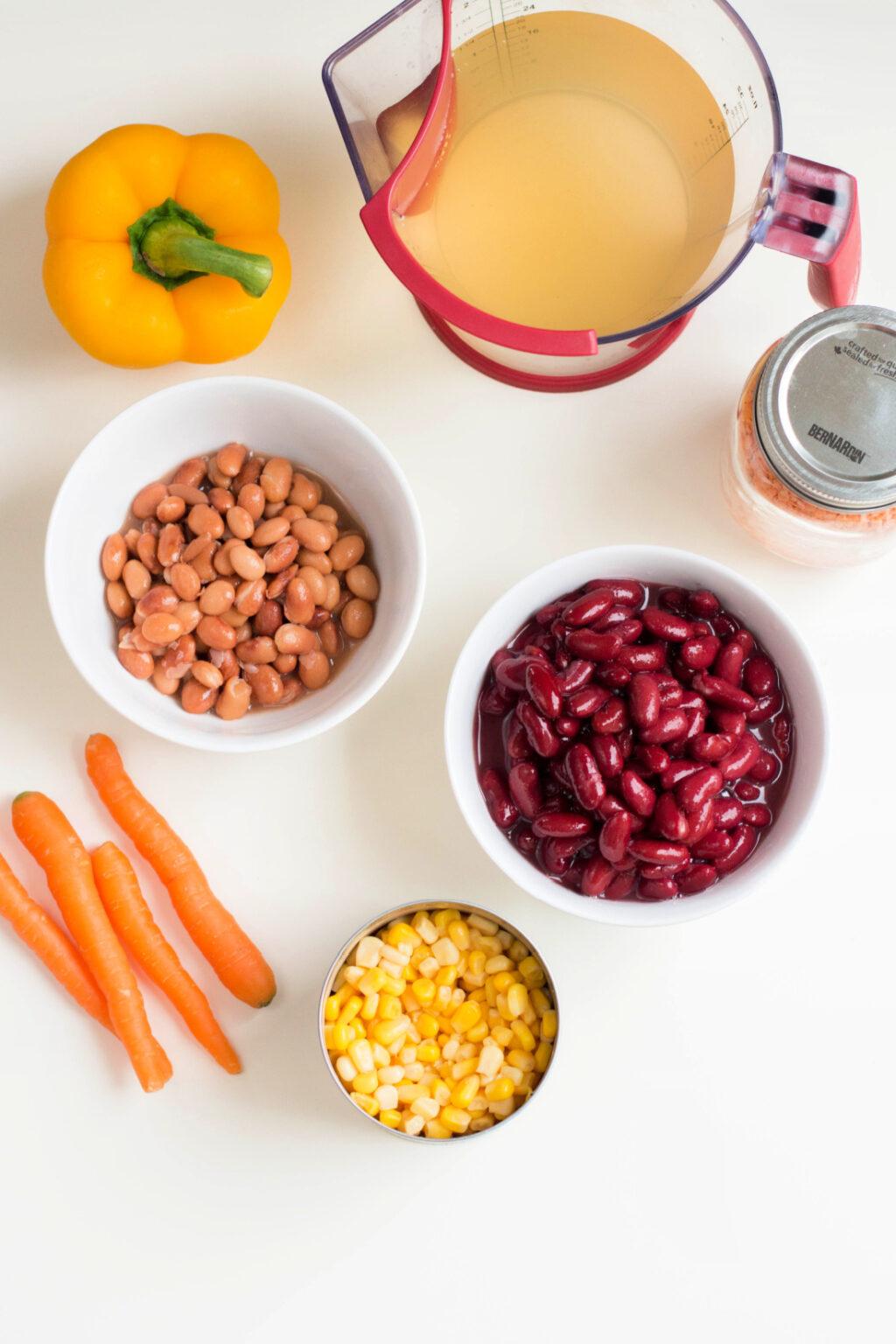 Various ingredients for cooking Instant Pot Vegetarian Chili displayed on a white surface, including bowls of beans, corn, carrots, a bell pepper, and a jug of liquid.