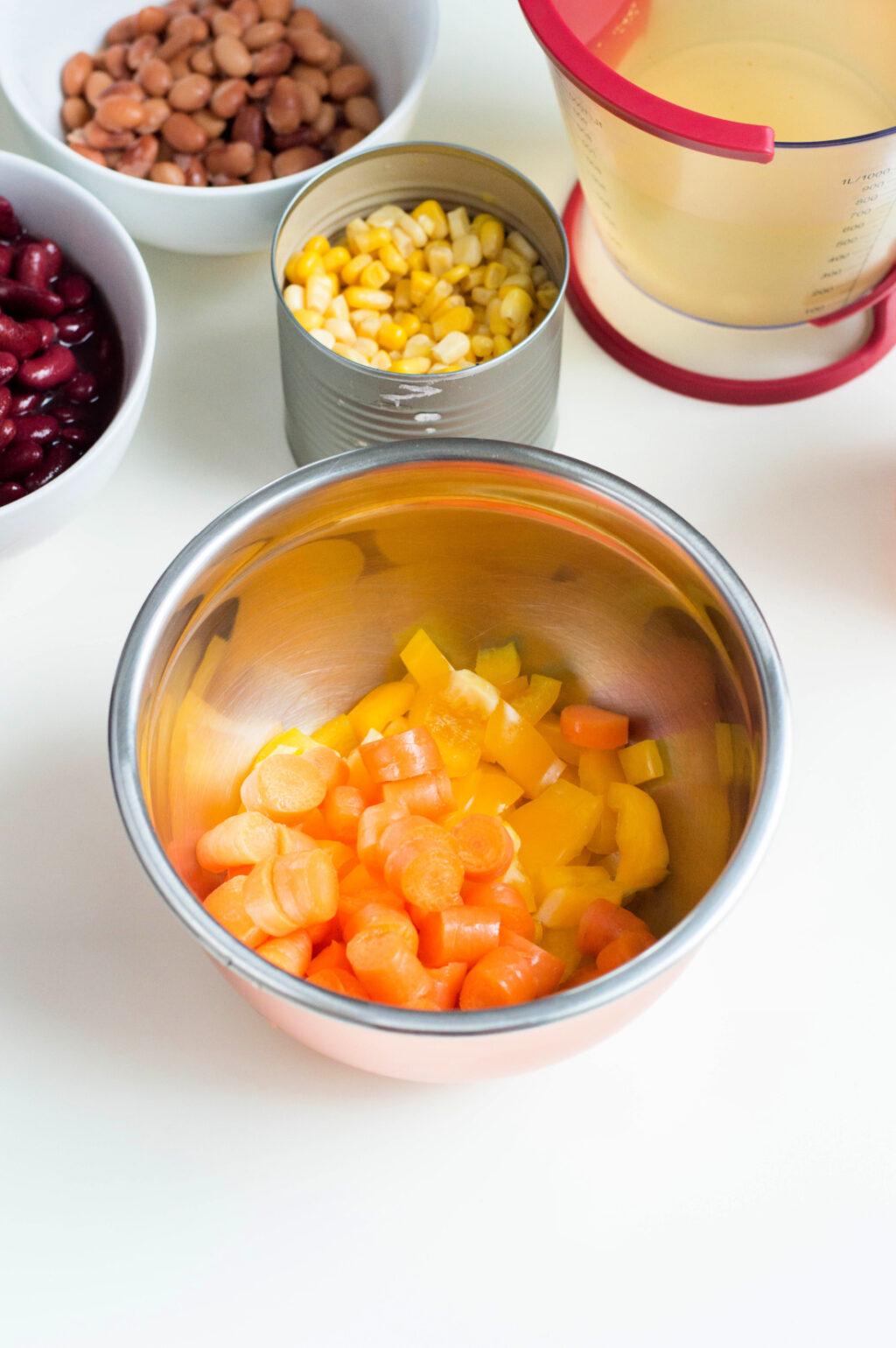 A metal bowl containing diced carrots and yellow bell peppers, with surrounding ingredients like corn, beans, and a measuring cup of liquid for a vegetarian instant pot chili.