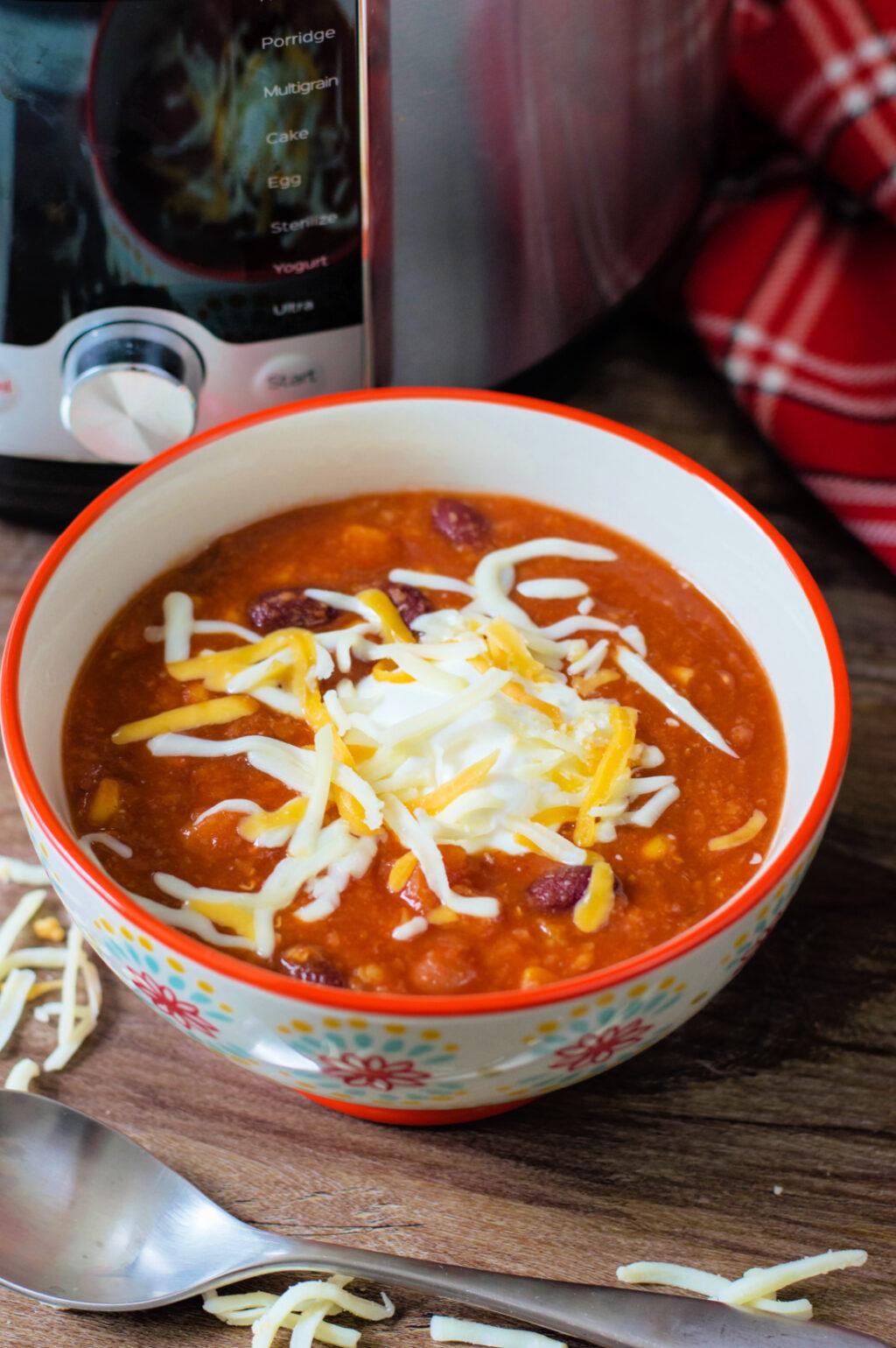 A bowl of vegetarian chili with beans and shredded cheese, placed on a wooden table next to a metal spoon and a red plaid cloth.