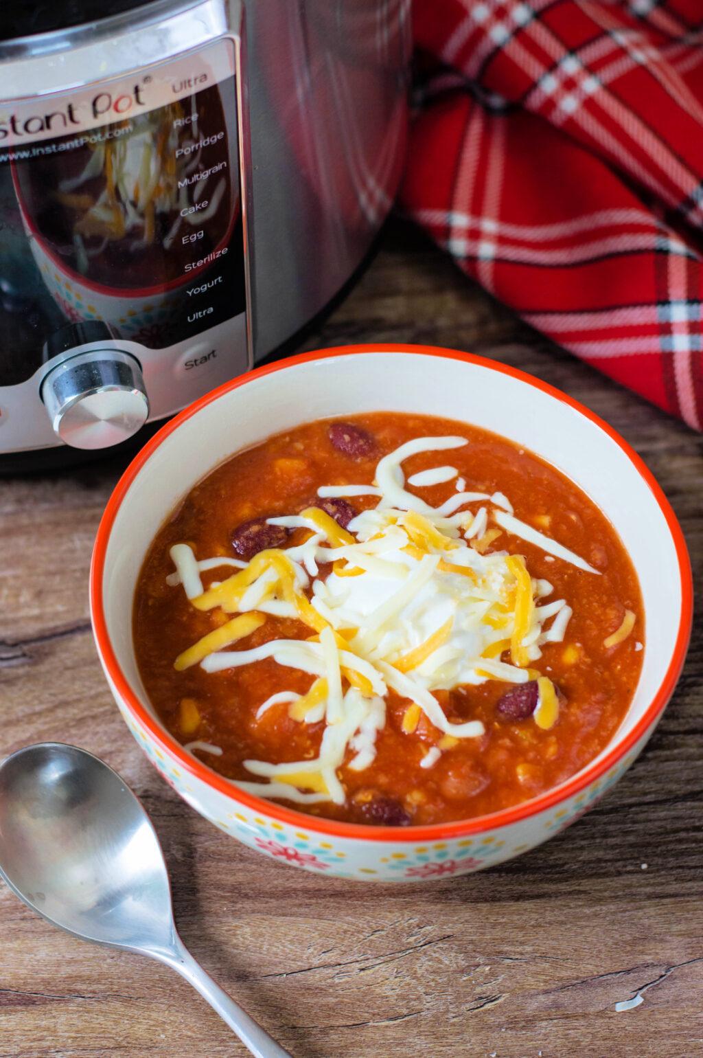 A bowl of vegetarian instant pot chili topped with shredded cheese and sour cream, next to an instant pot and a spoon, on a wooden surface.
