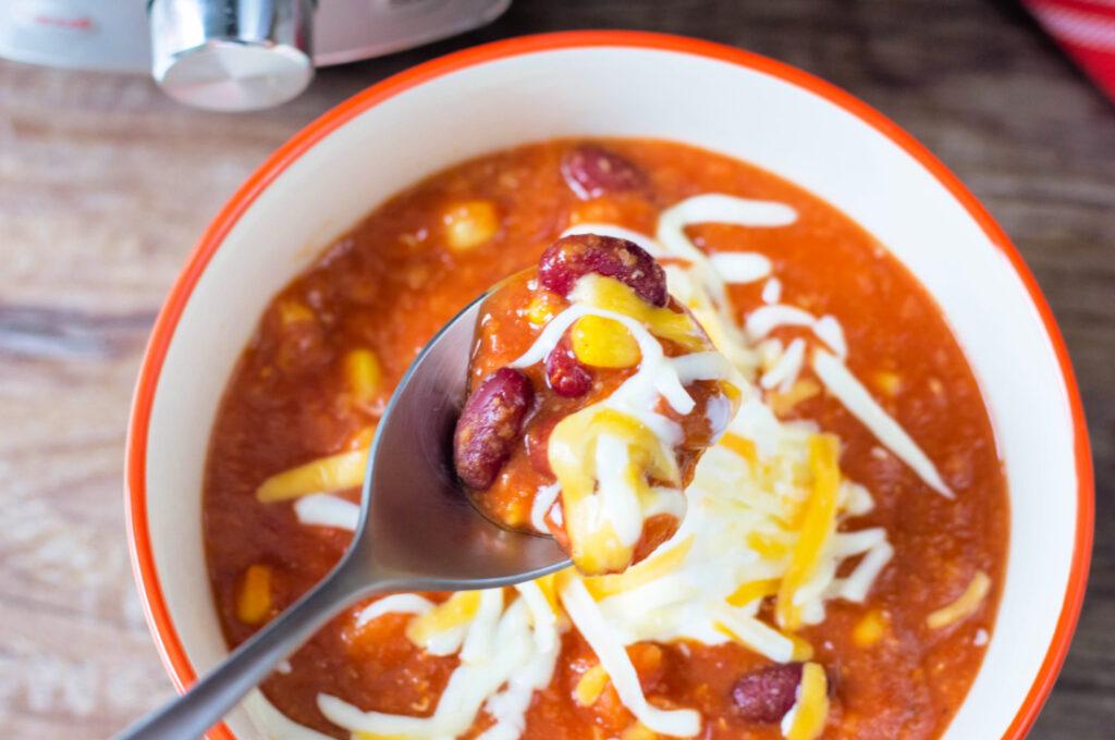 A bowl of vegetarian chili made in an Instant Pot, featuring tomatoes, corn, beans, and melted cheese, served with a spoon lifting some of the soup.