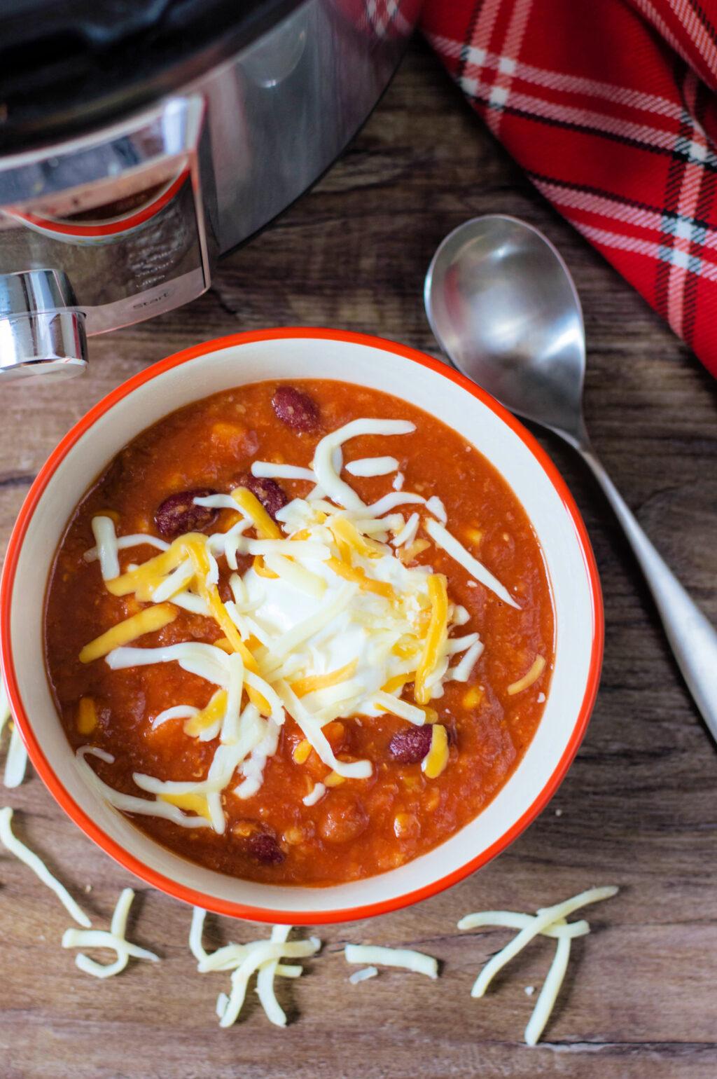 Bowl of Instant Pot Vegetarian Chili topped with shredded cheese and a dollop of sour cream, on a wooden table with a spoon and a checkered napkin on the side.